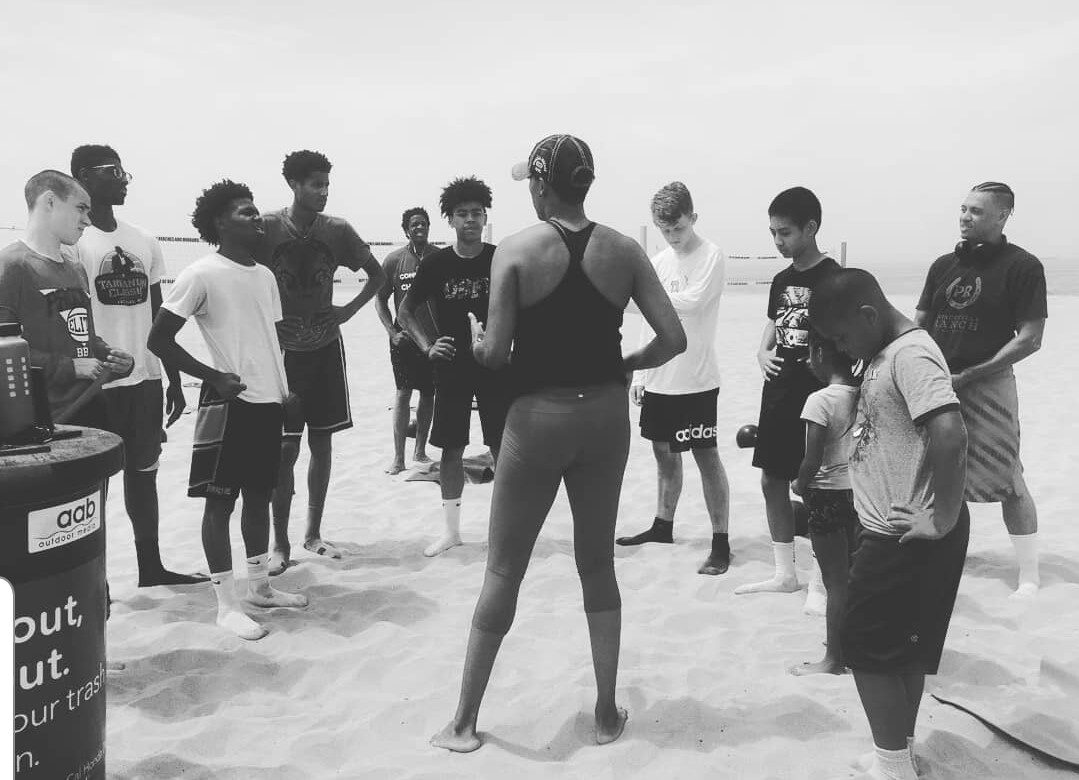 Coach Peters leading a team huddle with young athletes during a beach training session.